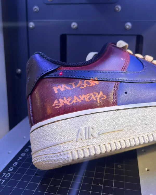 Une Air Force 1 bordeaux dans la machine de gravure laser de la boutique, l'inscription Maison Sneakers gravée sur le côté